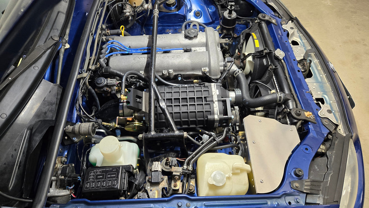 High three-quarter view of a 1999–2005 Miata engine bay with the supercharger and covered coldside intake
